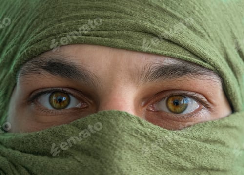 Preview: close up of the green eyes of a man whose face is covered with a blue bandage