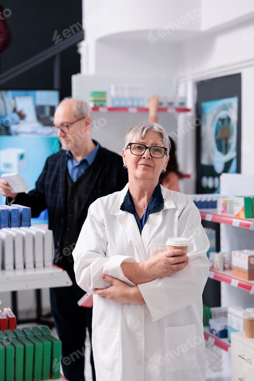Preview: Elderly drugstore worker in white coat holding cup of coffee looking at camera while working in