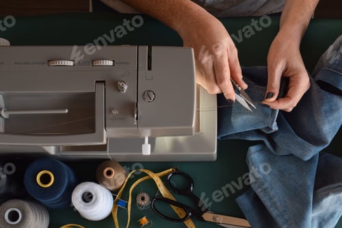 Preview: Seamstress cutting thread while using sewing machine to hem jeans pants top view, from above