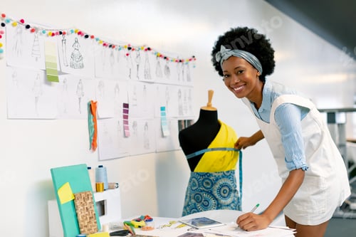 Preview: Happy African american female graphic designer writing on paper while measuring mannequin in office