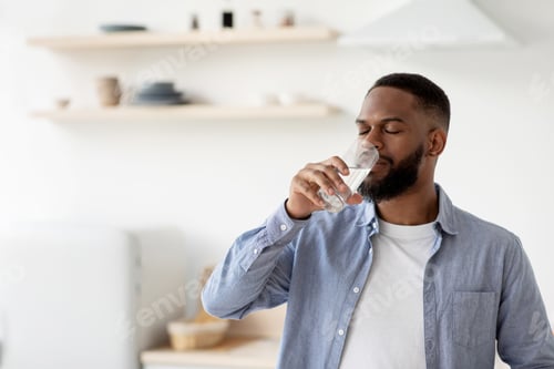 Preview: Calm young african american male with beard drinks of clean water from glass with closed eyes