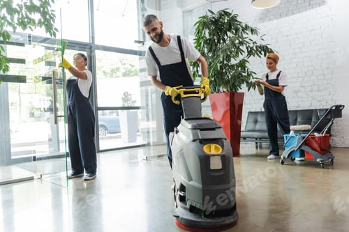 Preview: man in workwear with floor scrubber machine near interracial women cleaning office lobby