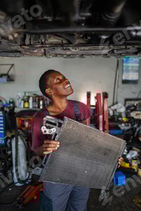 Preview: Young african female mechanic holding car radiator and looking up at vehicle in a garage
