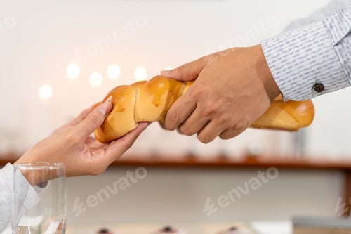 Preview: Two hands pass challah bread during a Hanukkah meal, with sufganiyot and Hanukkah