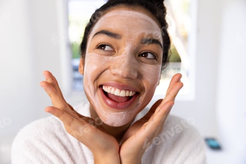 Preview: Portrait of happy biracial woman in bathrobe with face mask in sunny bathroom