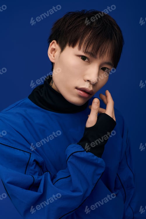 Preview: Captivating young Asian man showcasing charm against a bold blue backdrop while thoughtfully posing