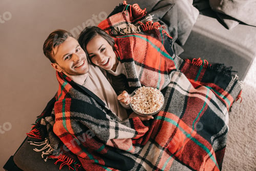 Preview: high angle view of young couple relaxing on couch with bowl of popcorn and covering with plaid