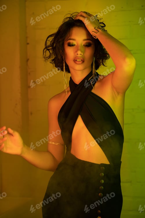 Preview: Sexy asian woman in dress touching hair while standing in smoke and neon light in night club
