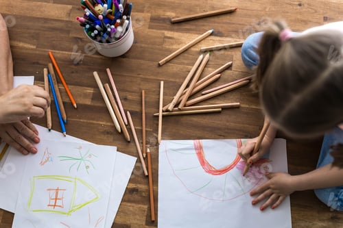 Preview: A 5-year-old girl happily draws at a table surrounded by a rainbow of colorful pencils,top view