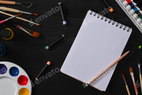 Preview: Sketchbook, paint brushes, tube paint, watercolor and palette on black wooden table.