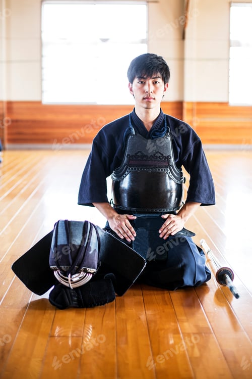 Preview: Male Japanese Kendo fighter kneeling on wooden floor, looking at camera.