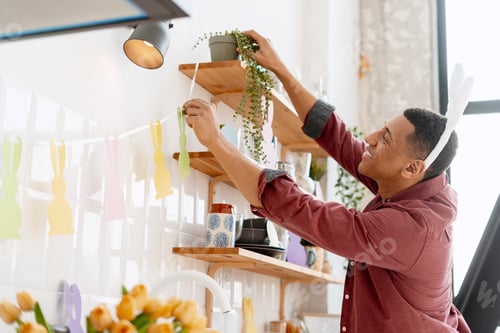 Preview: Smiling man hanging colorful bunny decorations, preparing kitchen for Easter celebration