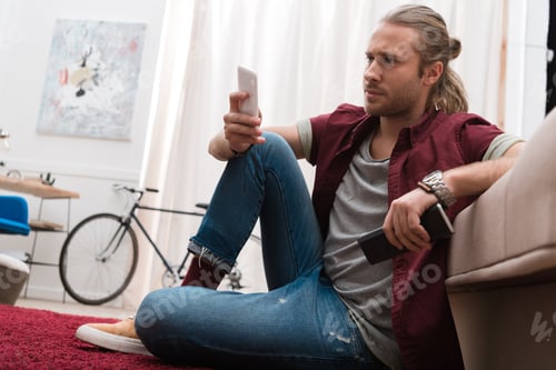 Preview: handsome man with remote control and smartphone at home
