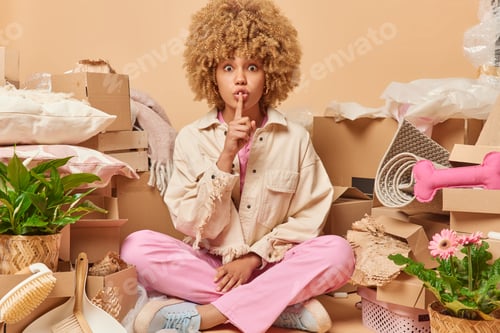 Preview: Indoor shot of curly haired woman sits in lotus pose makes silence gesture surrounded by carton