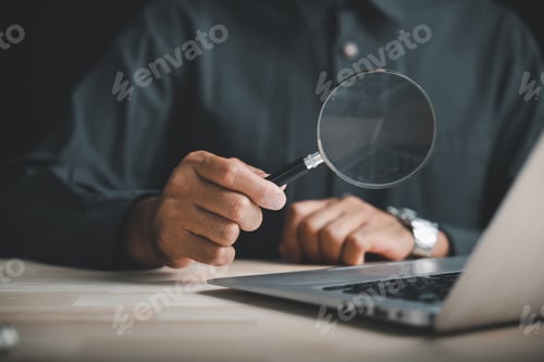 Preview: Closeup of a hand holding a magnifying glass over a laptop keyboard, searching for information