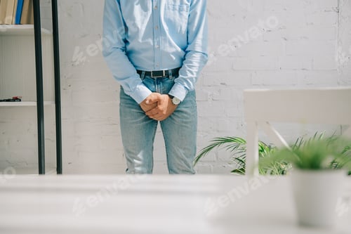Preview: cropped view of man holding clenched hands near groin area while standing by white wall