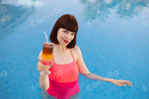 Preview: smiling pretty woman in pink swimwear, holding a fresh cold cocktail while swimming in the blue pool