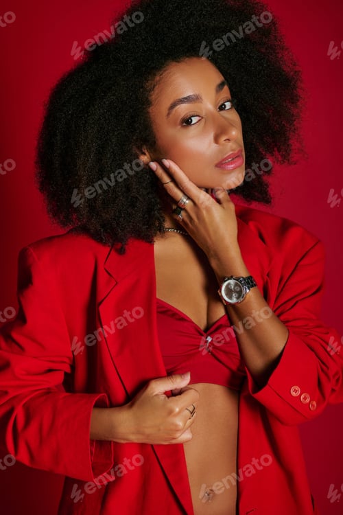 Preview: good looking alluring african american woman in vibrant attire looking at camera on red backdrop