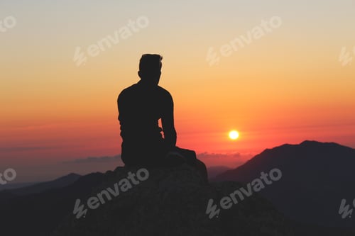 Preview: Silhouette of a male sitting on the rocks with a beautiful sunset scenery in the background