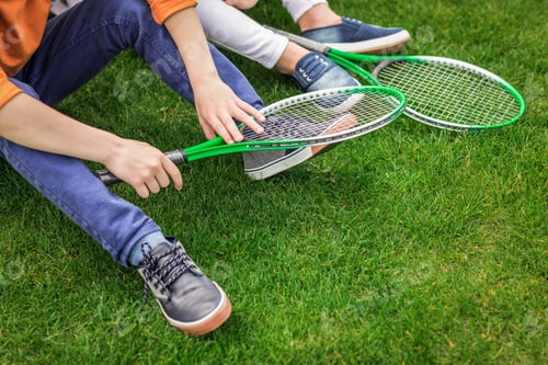 Preview: cropped shot of kids with badminton rackets sitting on green lawn