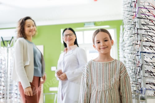 Preview: cheerful girl looking at camera near blurred mom and asian ophthalmologist in optics store