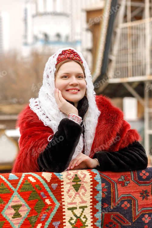 Preview: A Russian girl in red traditional Russian clothes and white shawl near wooden house in spring