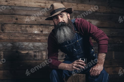 Preview: Vintage Worker Man With Long Gray Beard In Jeans Dungarees Holding Whiskey. Sitting On Wooden Crate