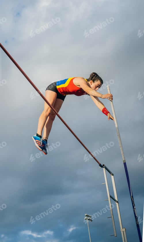 Preview: Female pole vaulter in mid-air over the bar against a cloudy sky.
