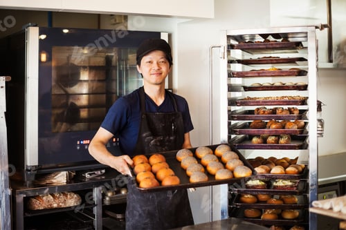 Preview: Man working in a bakery, holding large tray with freshly baked rolls, smiling at camera.