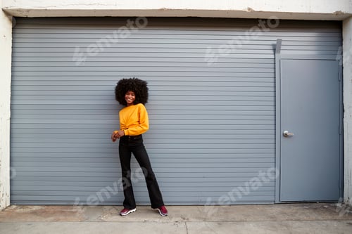 Preview: Hip young black woman standing against grey security shutters smiling to camera, full length
