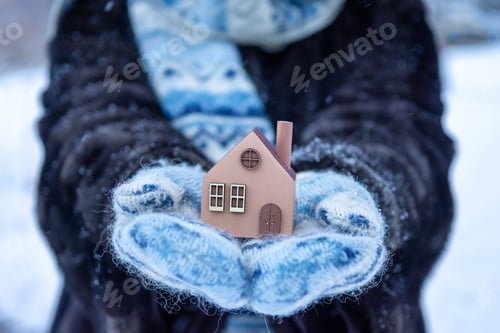 Preview: a woman in fluffy mittens holds a small house in her hands on a cold winter day.