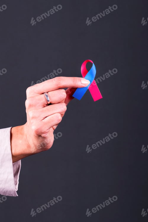 Preview: Woman’s hand holding a pink and blue ribbon, symbol of infant loss and pregnancy loss awareness