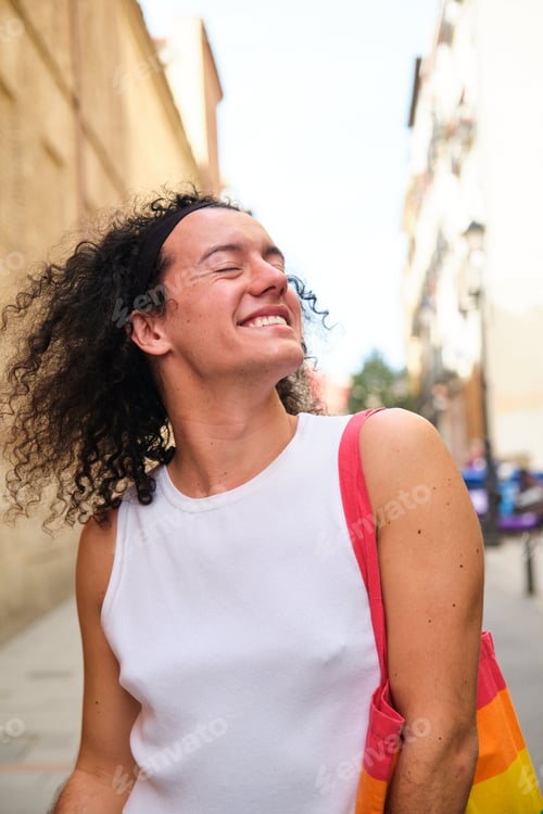 Preview: Gay man with curly hair is smiling and holding a rainbow bag.