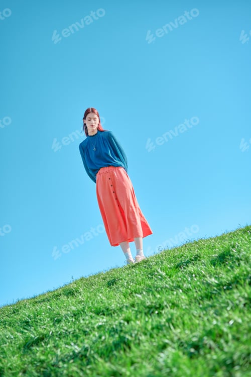 Preview: Teenage girl in a blue blouse and coral skirt standing on a grassy slope against a clear blue sky.