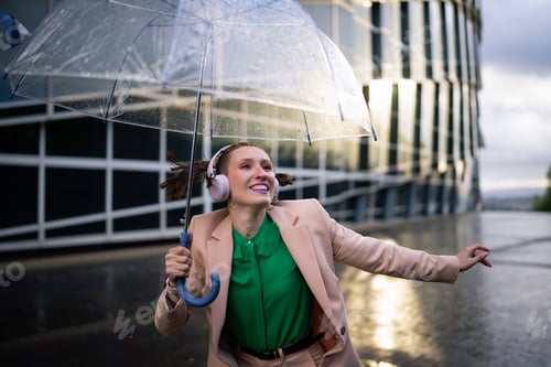 Preview: Happy businesswoman dancing in rain with transparent umbrella and headphones