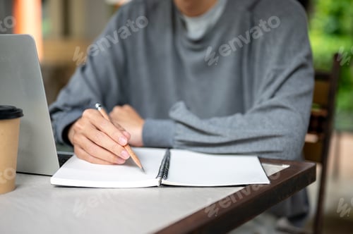 Preview: Close up of a man's hand holding pencil writing in notebook aside laptop and coffee at wooden table.