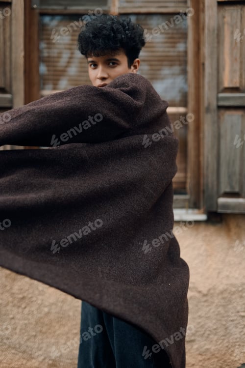 Preview: Fashionable young man in brown coat and scarf standing in front of window with arms outstretched
