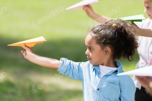 Preview: side view of little african american girl throwing paper plane while playing with friends