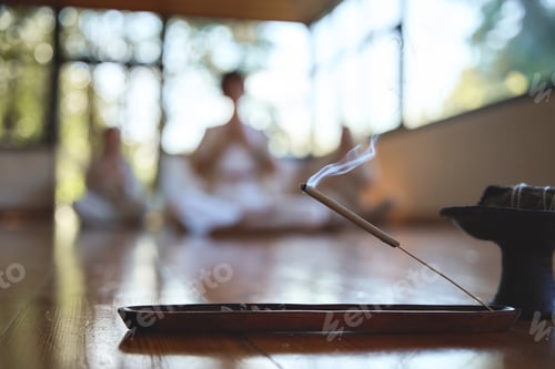 Preview: Incense stick with smoke on wooden holder with meditating people on background.