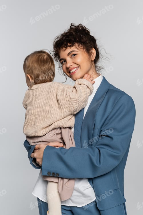 Preview: cheerful businesswoman in blue suit holding in arms baby girl isolated on grey