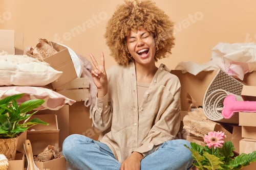 Preview: Joyful woman with curly hair dressed casually makes peace sign smiles happily glad to become owner o