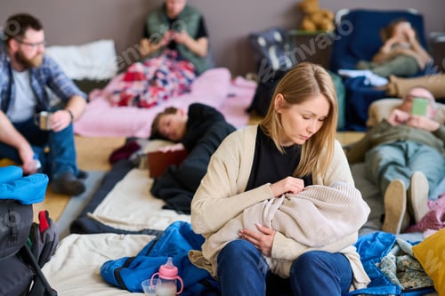 Preview: Young blond female refugee lulling baby wrapped into warm plaid on bed