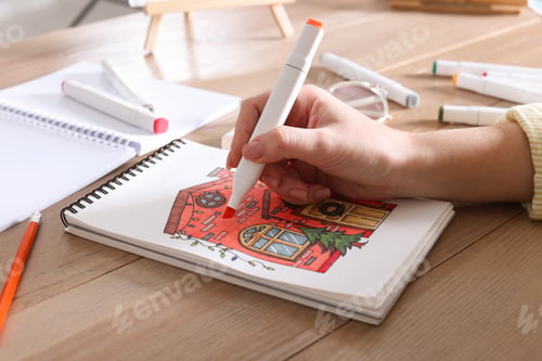 Preview: Woman drawing in sketchbook with felt tip pen at wooden table, closeup