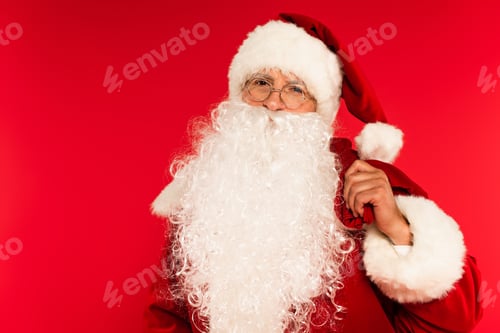 Preview: Bearded santa claus holding sack and looking at camera isolated on red