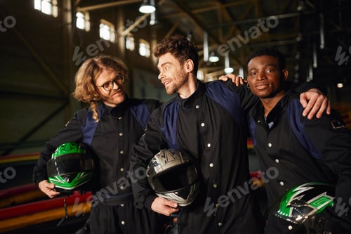 Preview: group of multicultural and happy go kart racers in protective suits hugging and holding helmets