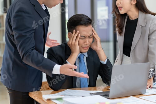 Preview: Stressed businessman holding head while coworkers argue at office desk with laptop