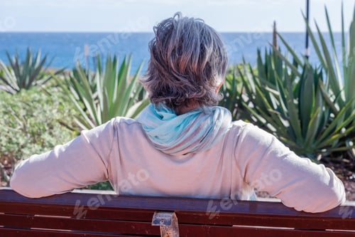 Preview: Rear view of an aged grey-haired woman relaxing in front to the sea, sitting on a bench looking away