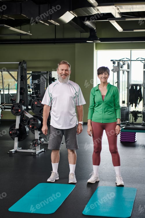 Preview: happy elderly couple standing near fitness mats, looking at camera, full length, active seniors