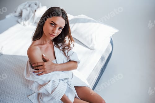 Preview: Portrait of beautiful brunette in white clothes that sits on the bed in room. Nice lighting