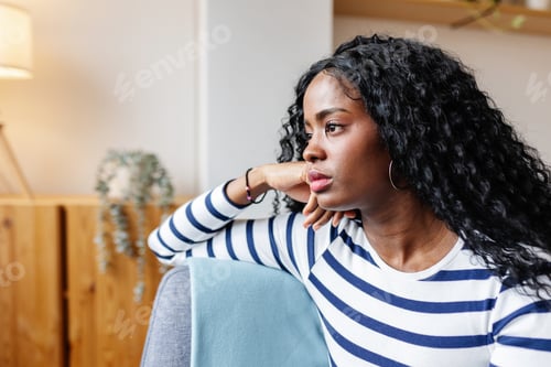 Preview: Sad young African American woman sitting on sofa looking away at home